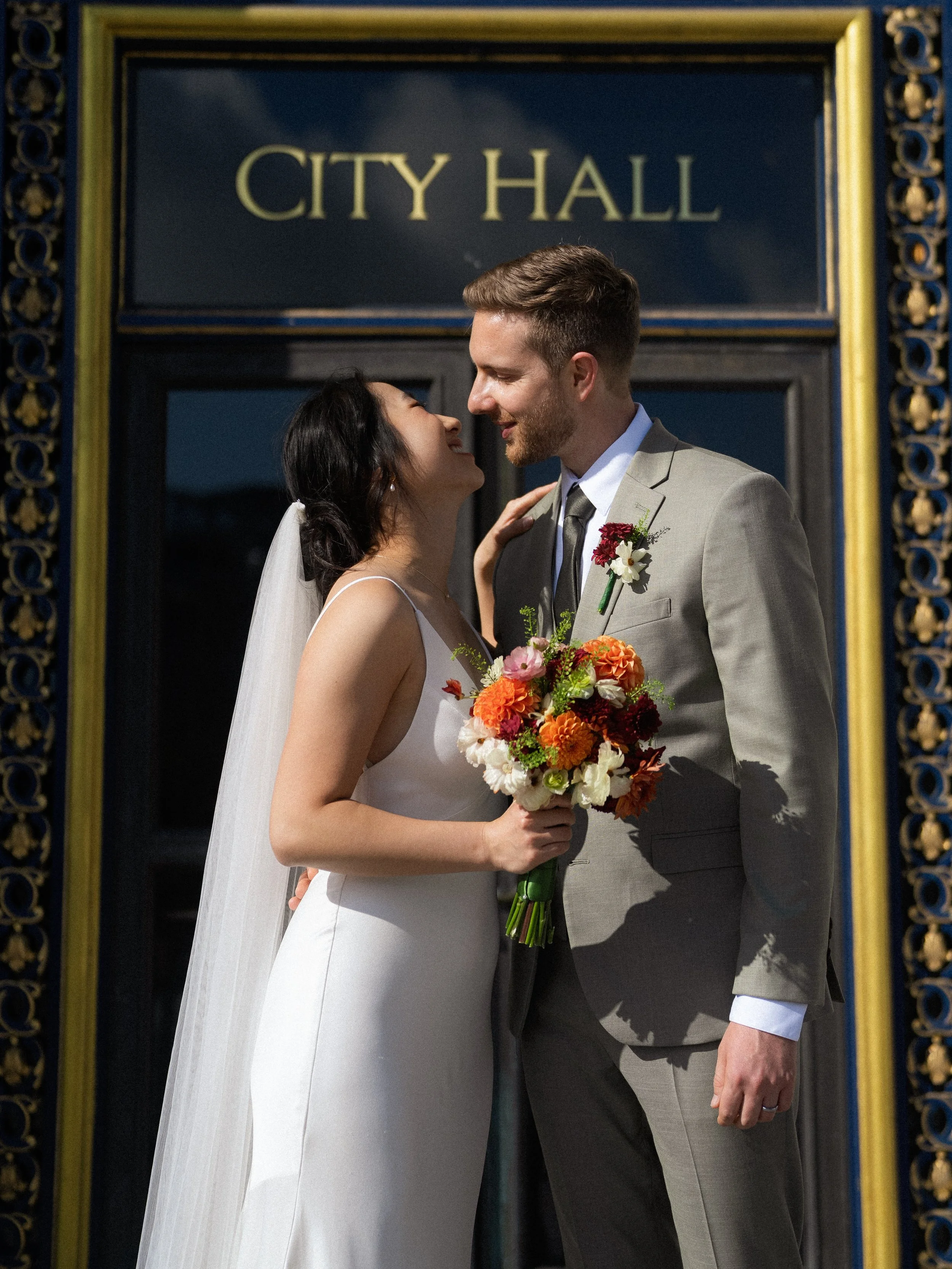 A bride and groom standing close together in front of a city hall sign, smiling and looking into each other's eyes. The bride holds a colorful bouquet of flowers, and both are dressed in formal wedding attire.