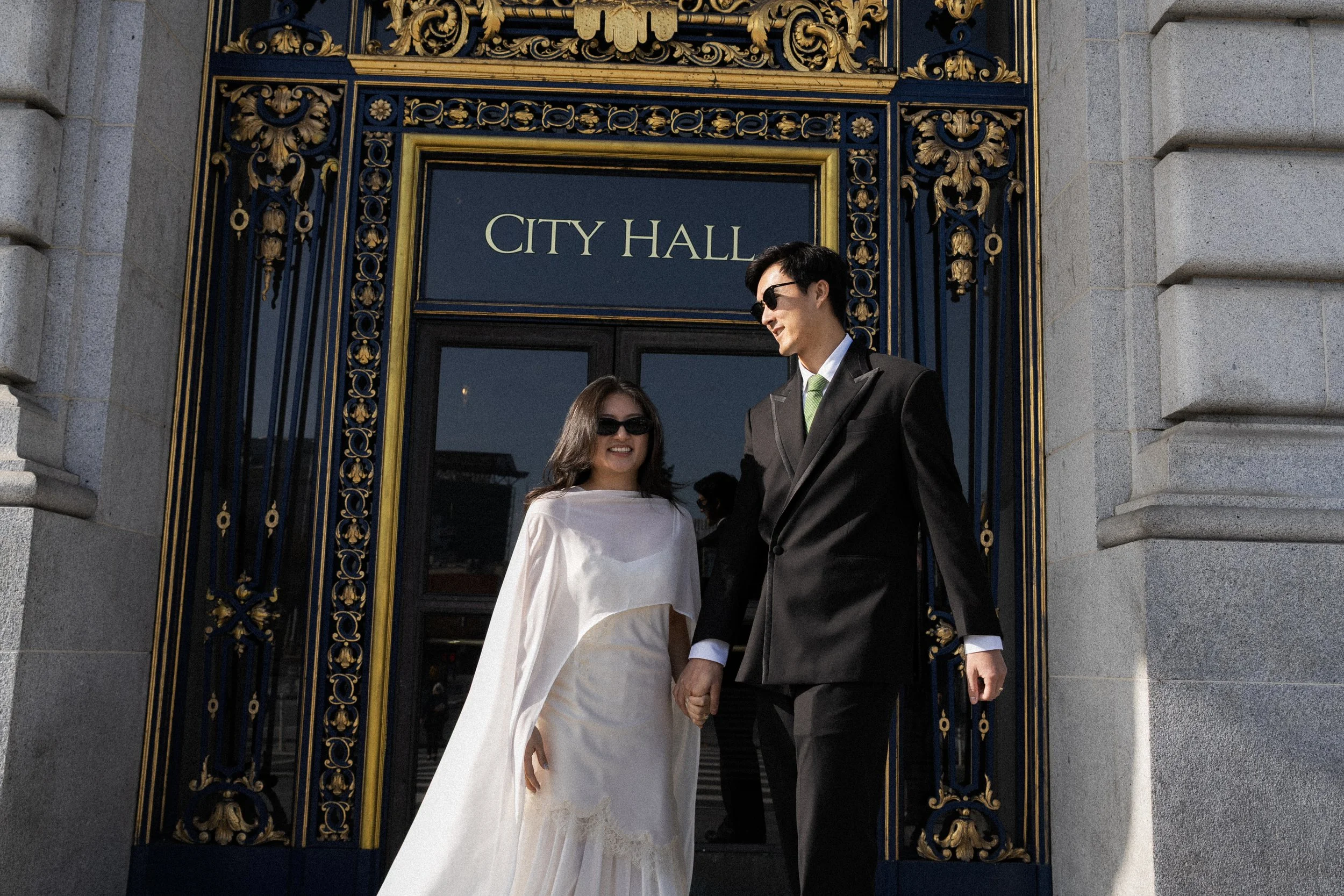 A couple, dressed in wedding attire, holding hands and smiling, standing in front of a city hall building with ornate gold and navy blue decoration above the door.