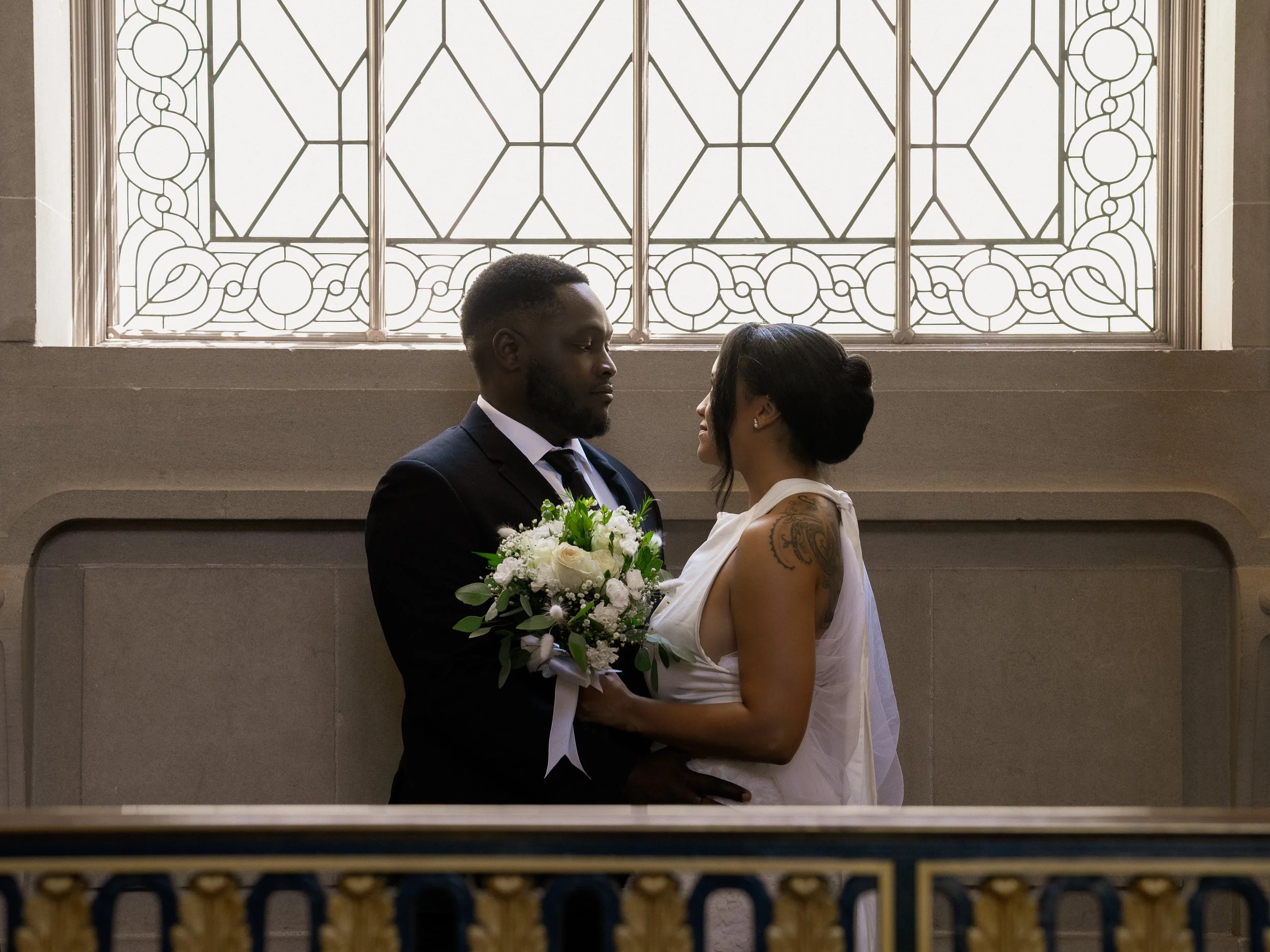 A bride and groom standing close together in a church, facing each other, with the bride holding a bouquet of white flowers, and a large decorative window in the background.
