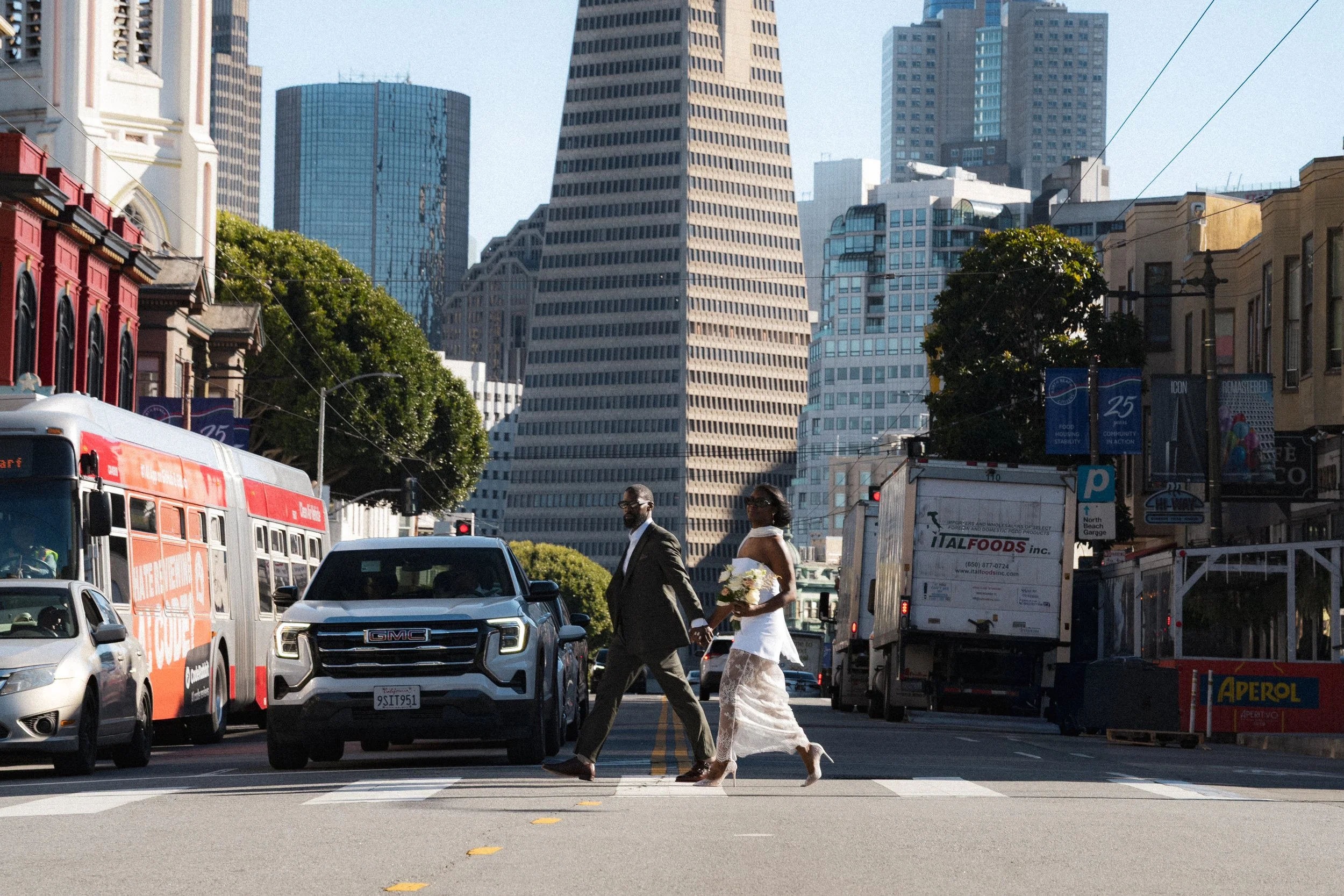 A city street scene with a couple holding hands crossing a crosswalk during daytime. The man is dressed in a suit, and the woman is wearing a white dress and high heels, holding a bouquet of flowers. There are various vehicles on the street, including a bus, cars, and trucks, with tall modern skyscrapers in the background.