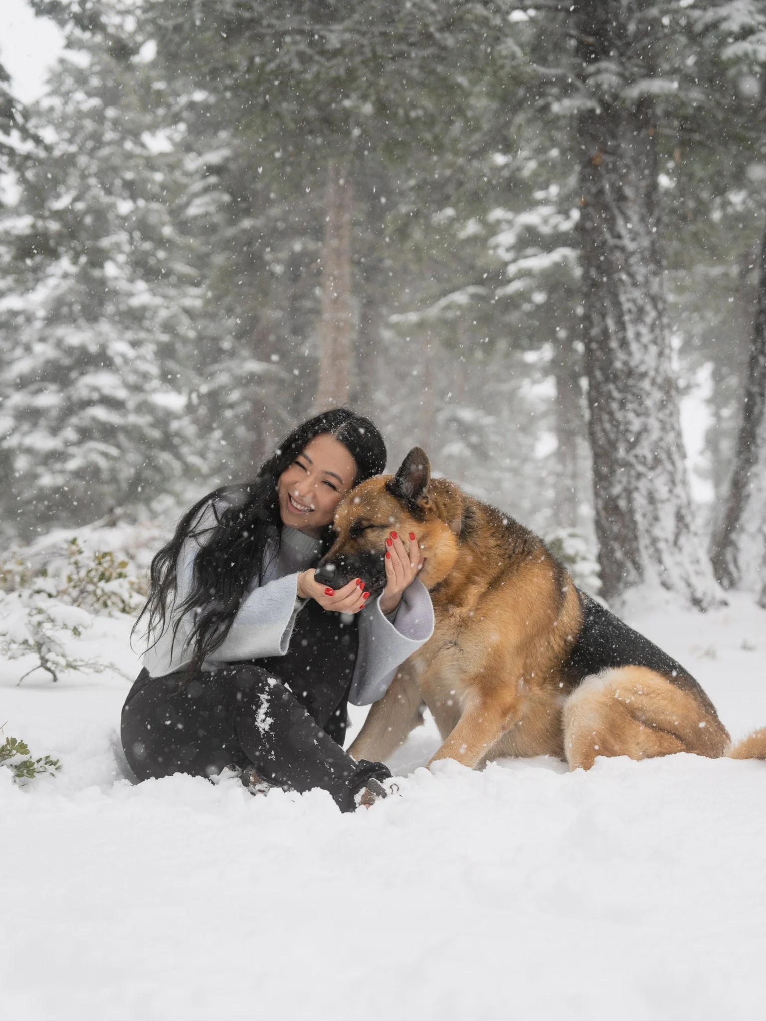 A woman with long black hair smiling and sitting in snow, hugging a large German Shepherd dog, in a snowy forest.