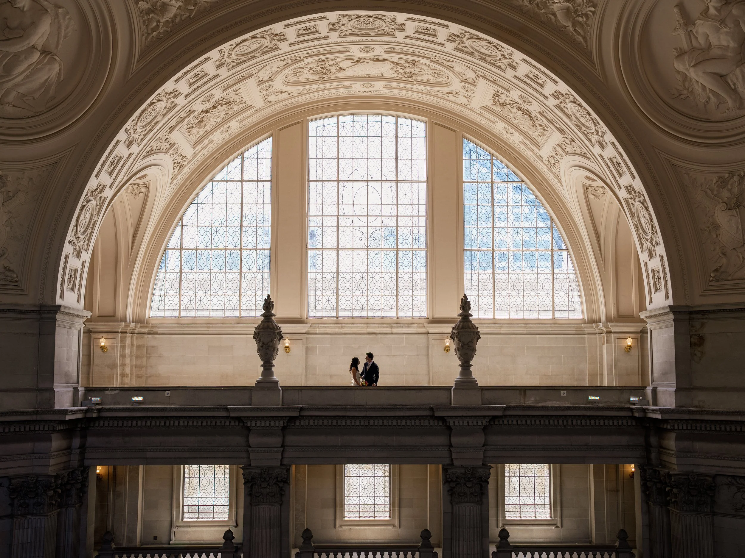 Two people in formal attire sharing a kiss inside a grand, historic building with ornate architectural details and large decorative windows.