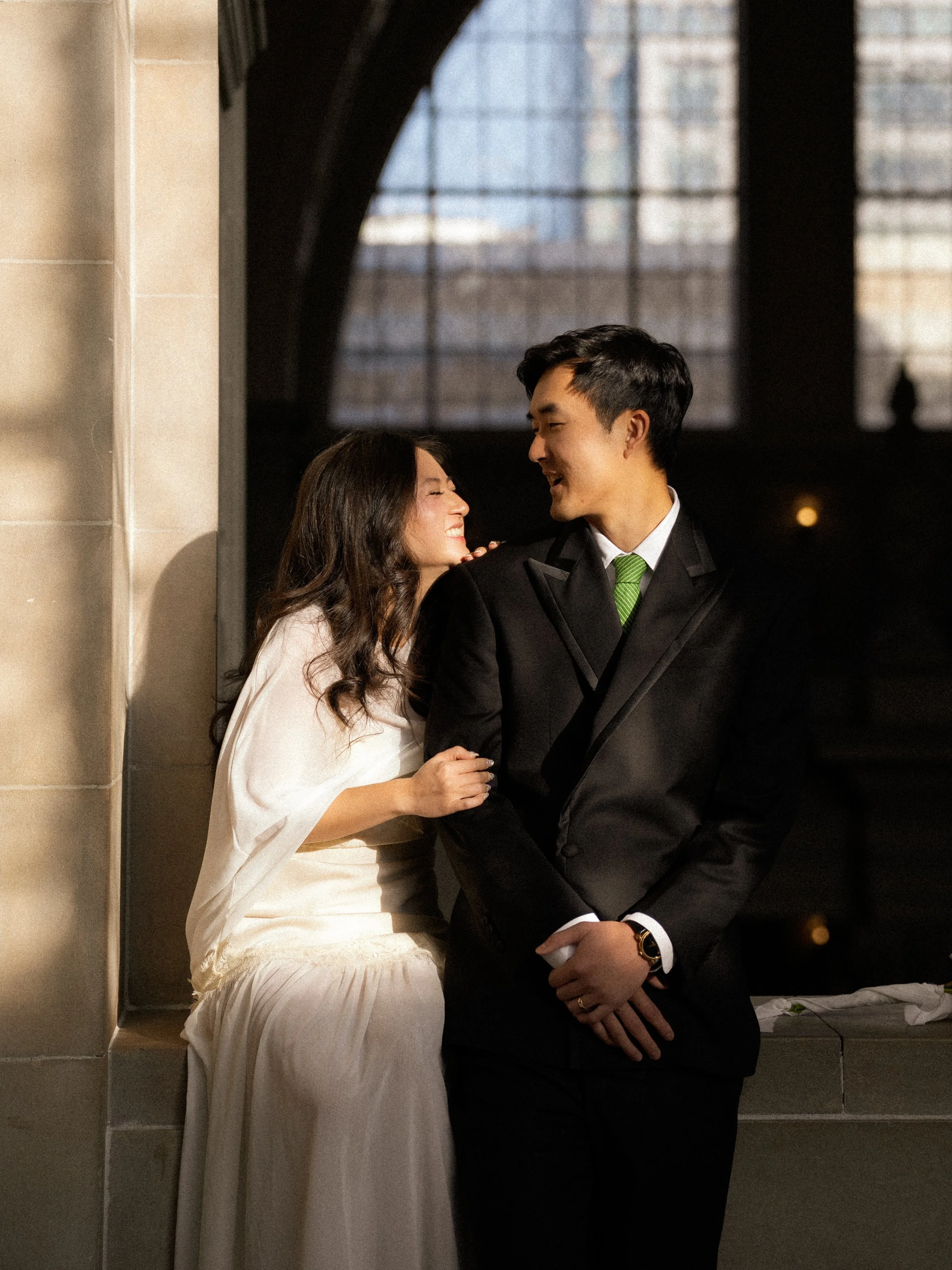 A couple dressed in formal attire, smiling and looking at each other, standing closely in front of a large window with a cityscape in the background.