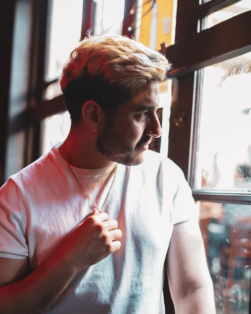 A man with styled blonde hair and a nose ring looking out a window in a cafe or restaurant, wearing a white t-shirt and a gold necklace.