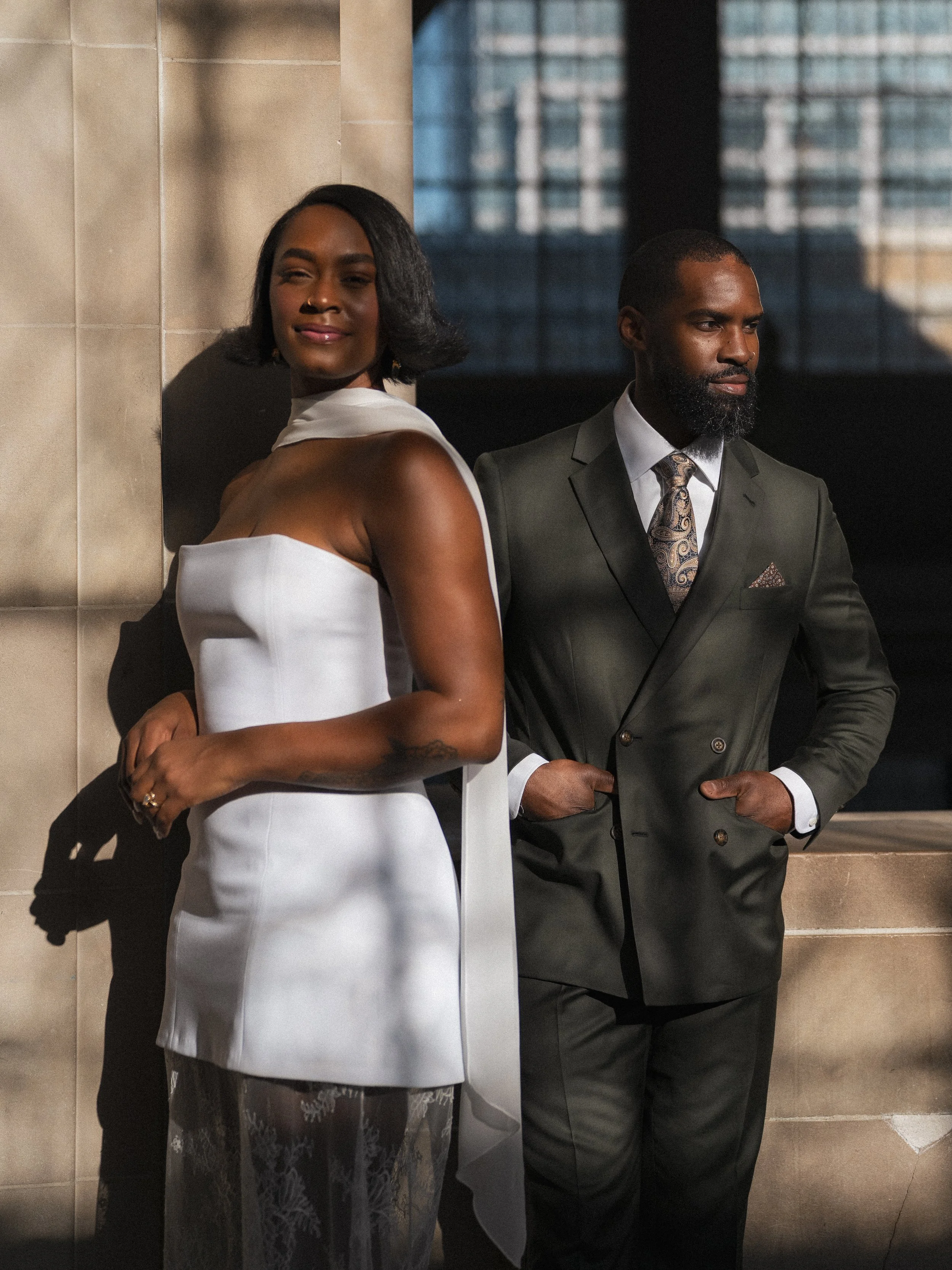 Two well-dressed individuals, a woman in a white dress and a man in a gray suit, standing in front of a cityscape through a window. The woman is leaning against a wall, smiling, and the man is standing beside her, looking to the side.