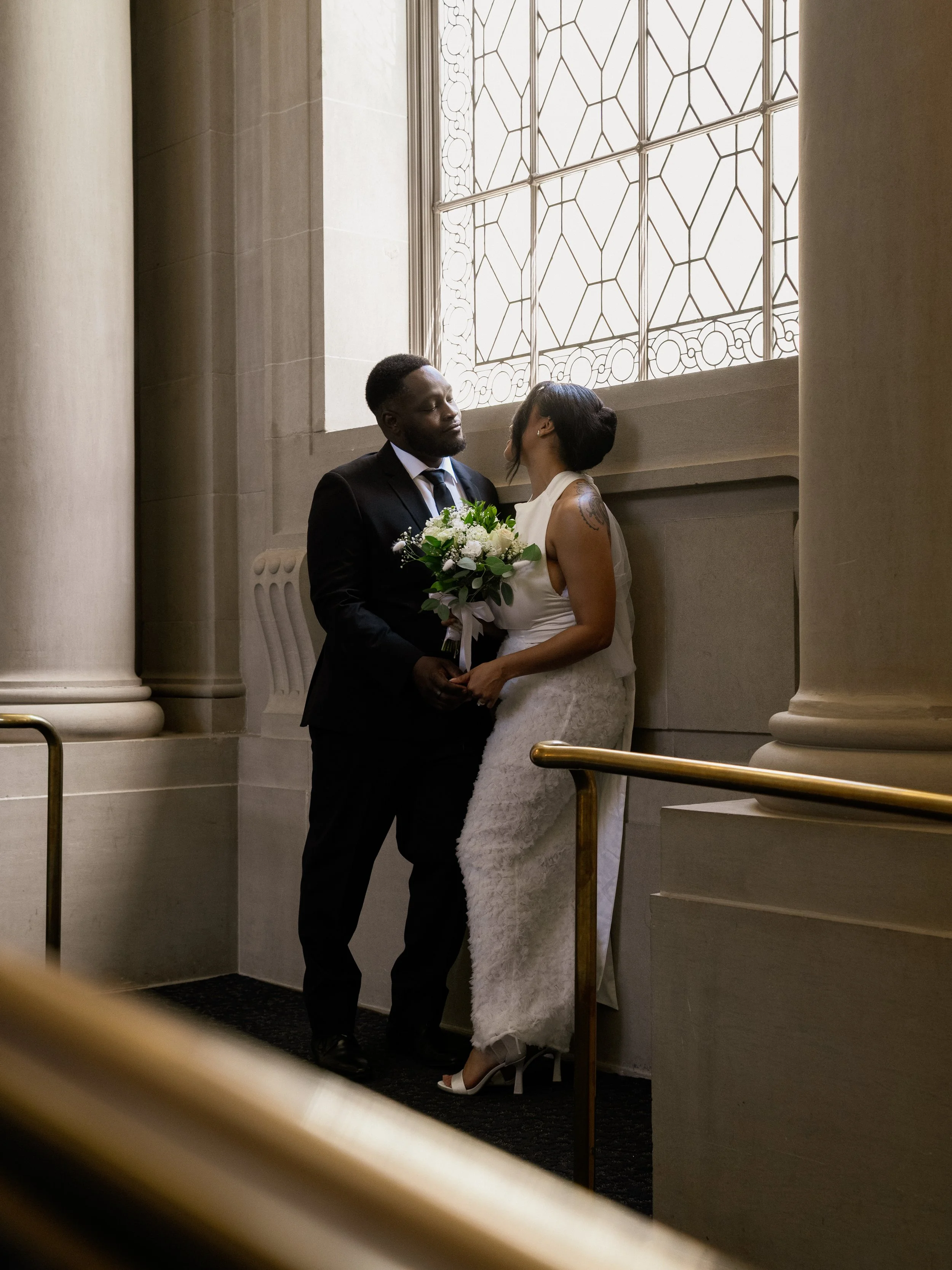 A newlywed couple sharing a romantic moment by a large stained glass window in a church. The groom holds a bouquet, and they are gazing into each other's eyes.