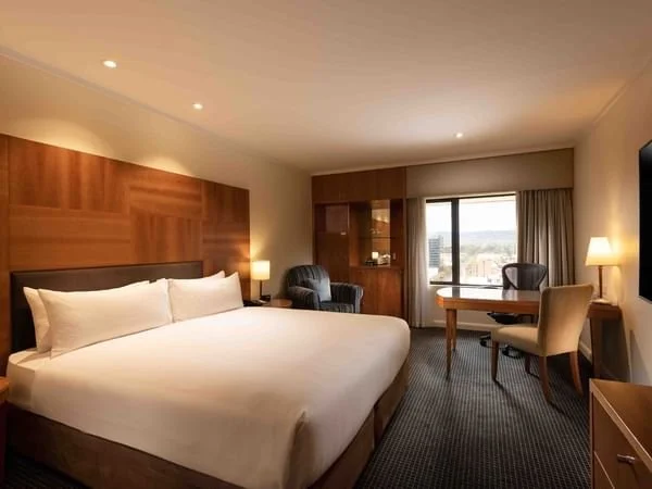 Modern hotel room with king-sized bed, wooden headboard, two lamps, a sitting area with two chairs and a desk near a window.