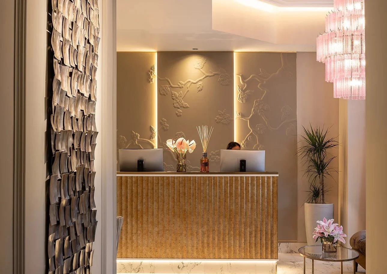 Hotel reception desk with two computer monitors, flowers, and decorative wall art behind. Soft lighting, pink chandelier, plant, and pink flowers on a side table.