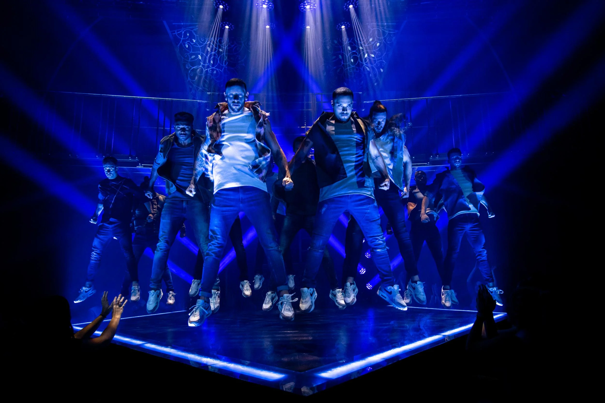 Magic Mike Live - A group of dancers performing on a stage illuminated with blue and purple lights, with some audience members raising their hands.