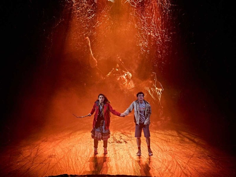 Two children holding hands on a stage with a fiery, volcanic or lava-like background.