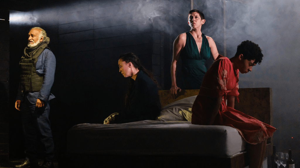 Four people in a dimly lit room, one elderly man standing on the left, three women sitting on or near a bed, with one of the women sitting on the edge of the bed and another standing behind it, in a scene that appears tense or dramatic.