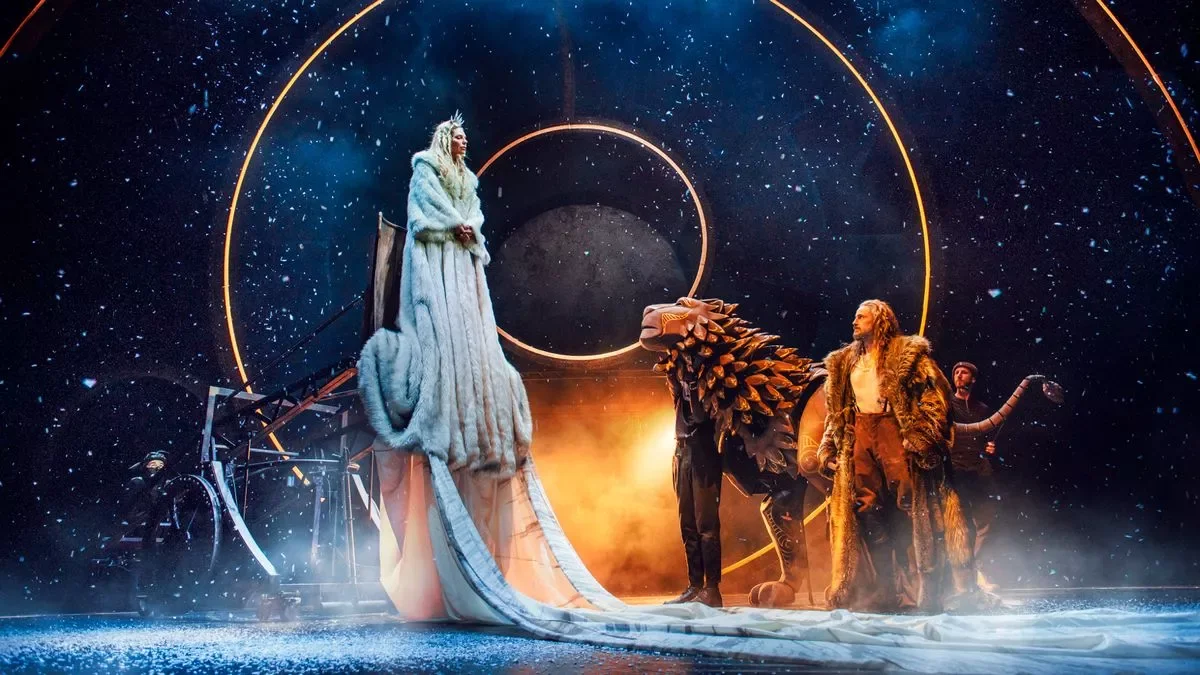 The image shows a theatrical stage performance with a winter fantasy theme. There is a woman dressed in a long, white fur coat with a crown, standing on a platform. Around her, there are performers in costumes resembling a dragon or mythical creatures. The background is designed to look like outer space with dark blue and starry patterns, and there are glowing circular lights. The scene is illuminated with warm, orange lighting, creating a mystical atmosphere.