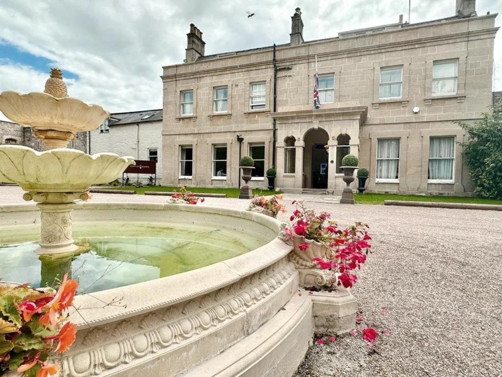 A grand stone building with a symmetrical facade, four windows on each floor, and an arched entrance with steps. British flag flying in front and bushes on either side. A decorative stone fountain with flowers is in the foreground. The sky is partly cloudy.