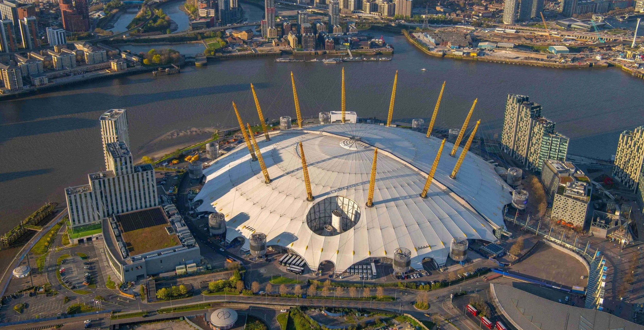 Aerial view of a large, modern sports arena with a white, tent-like roof and yellow support beams, surrounded by cityscape, river, and roads in a metropolitan area.