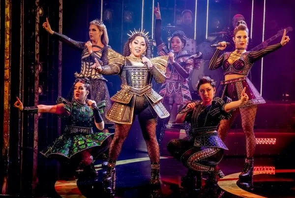 Eight women performing a stage dance. They are dressed in colorful, ornate costumes with metallic and studded details, and some wear crowns or headpieces. The background has stage lights and a dark backdrop, creating a vibrant theatrical atmosphere.