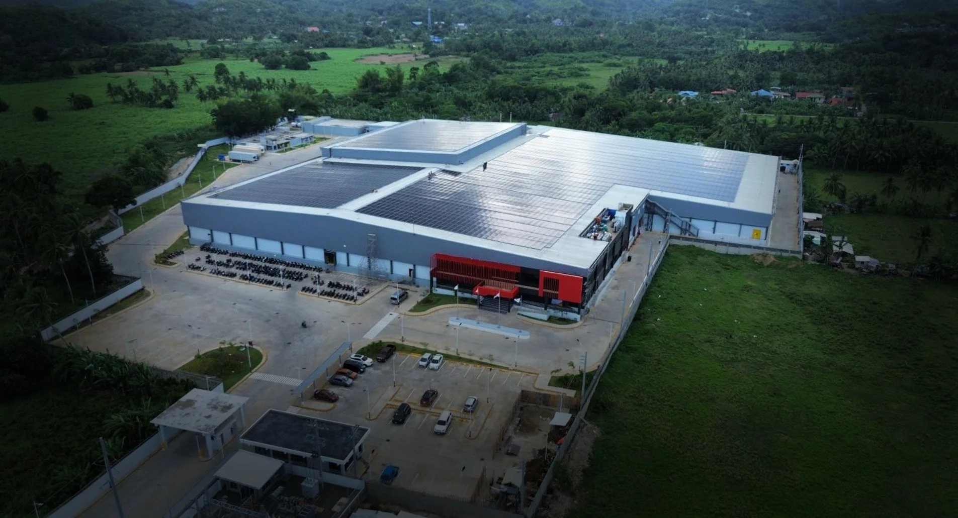 Aerial view of a large building with solar panels on the roof, surrounded by green fields and trees, with a parking lot and road in the foreground.