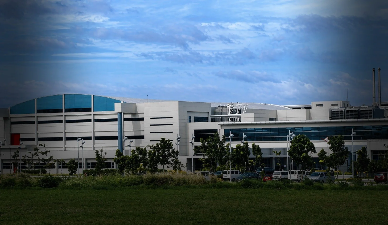 Modern white building with blue arched roof, large windows, surrounded by trees and a parking lot under a cloudy sky.