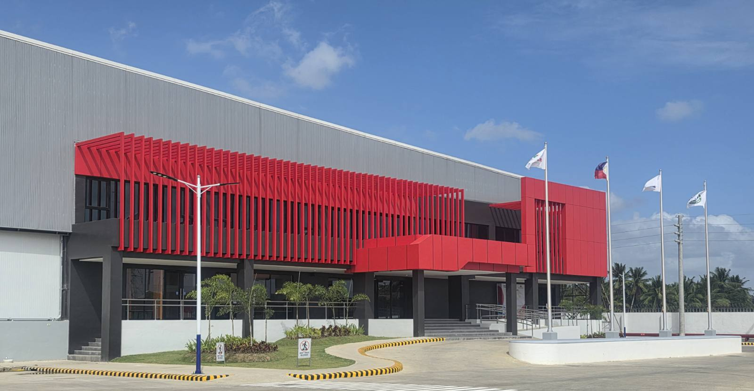 Modern commercial building with red and gray exterior, flagpoles with flags, trees, and a parking area under a blue sky.