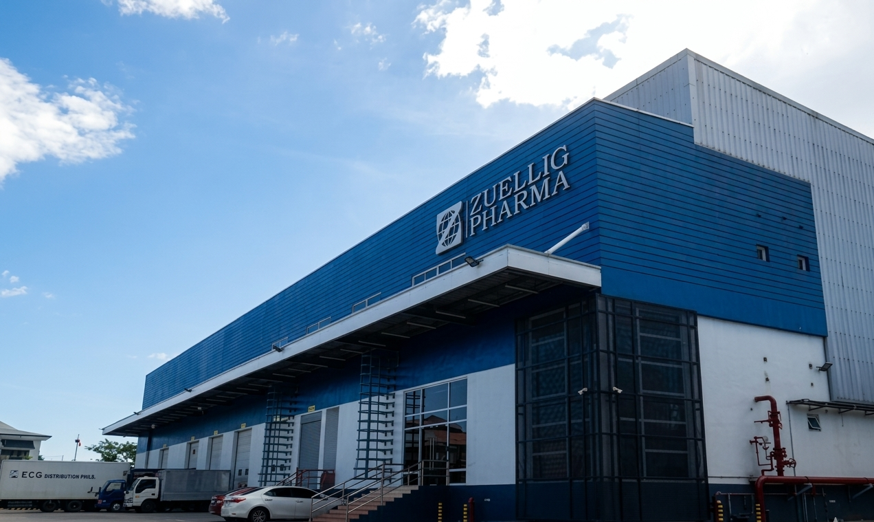 Blue and white exterior of a pharmaceutical building labeled "ZUELLIG PHARMA" with trucks and cars parked in front.