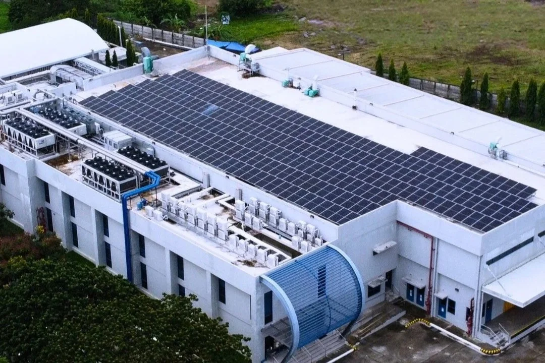 A building with a large rooftop covered in solar panels, with HVAC units and machinery on the side, surrounded by greenery and a fenced yard.