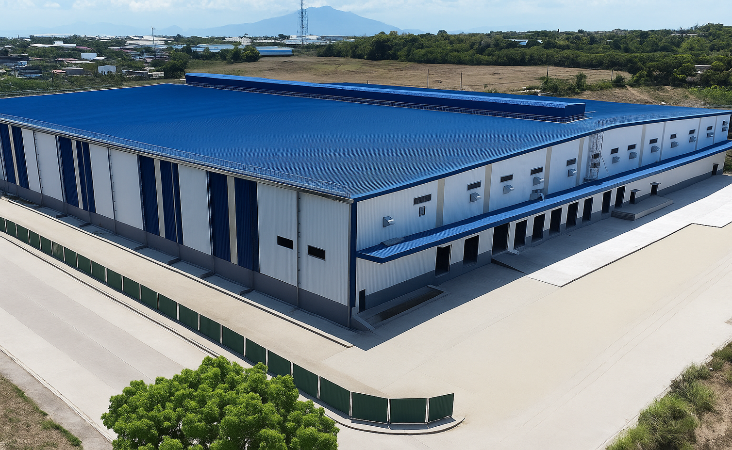 A large industrial building with a blue metal roof, white and blue exterior walls, and multiple loading doors, surrounded by a concrete yard and green fencing, set in a semi-rural area.