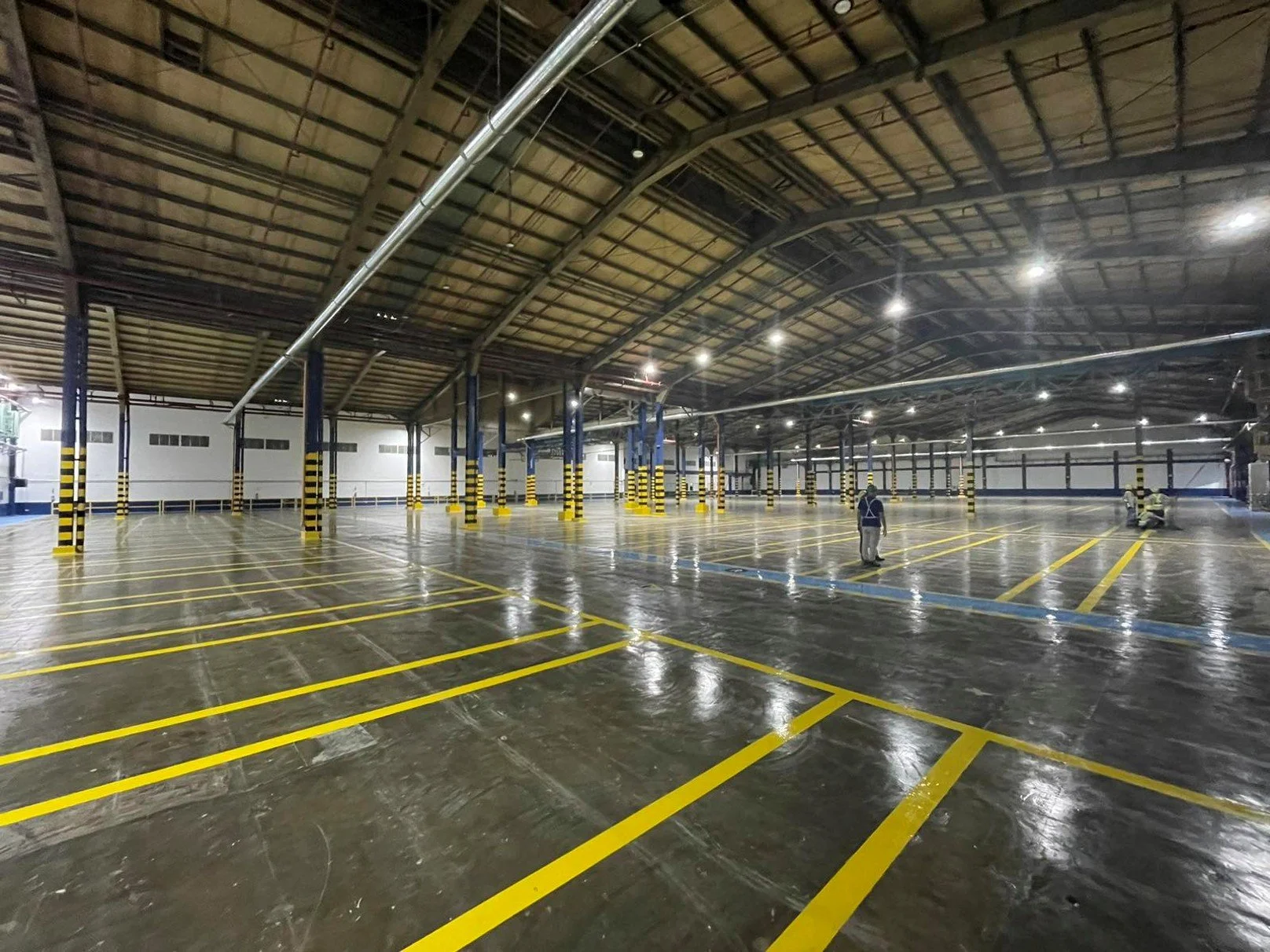 Empty indoor parking garage with yellow parking lines and black and yellow striped pillars, two workers in reflective vests working on the floor, and overhead lighting.