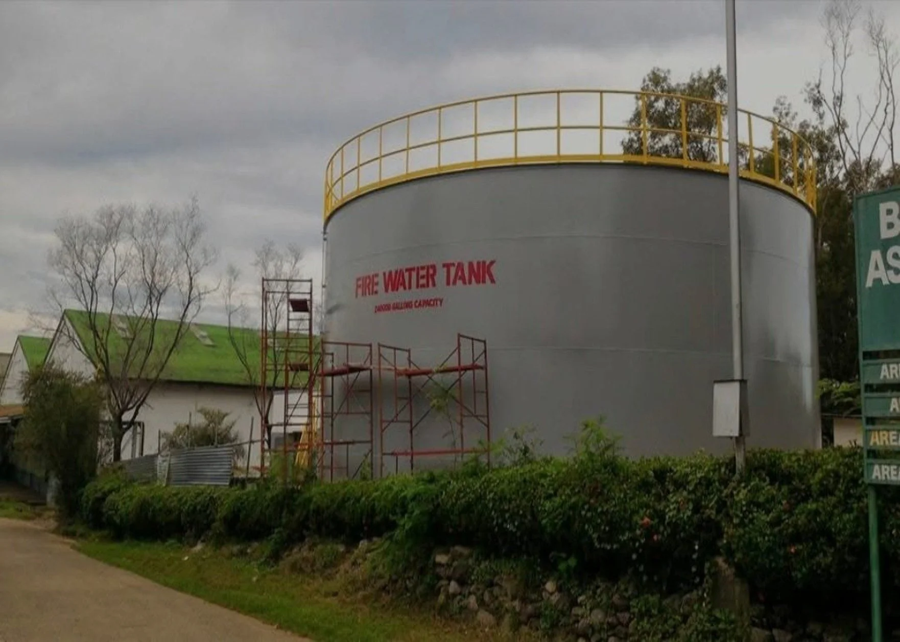 Large gray fire water tank with yellow safety railing on top, surrounded by some trees and buildings, and a cloudy sky in the background.