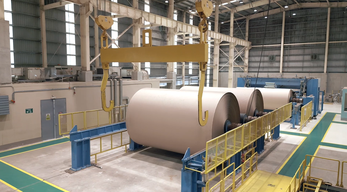 Large industrial setting with massive paper rolls on rollers, yellow overhead crane with hooks, and safety railings in a factory or warehouse.