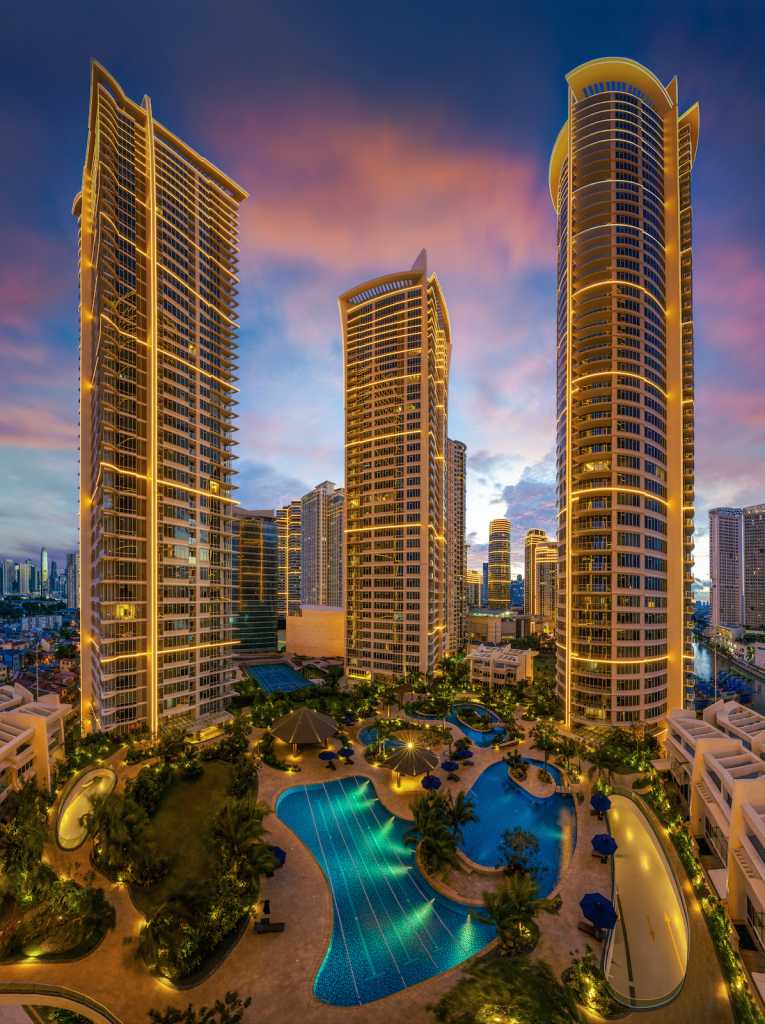 Night view of three high-rise buildings with illuminated edges surrounding a swimming pool area with umbrellas and lounge chairs, with a colorful sunset sky in the background.