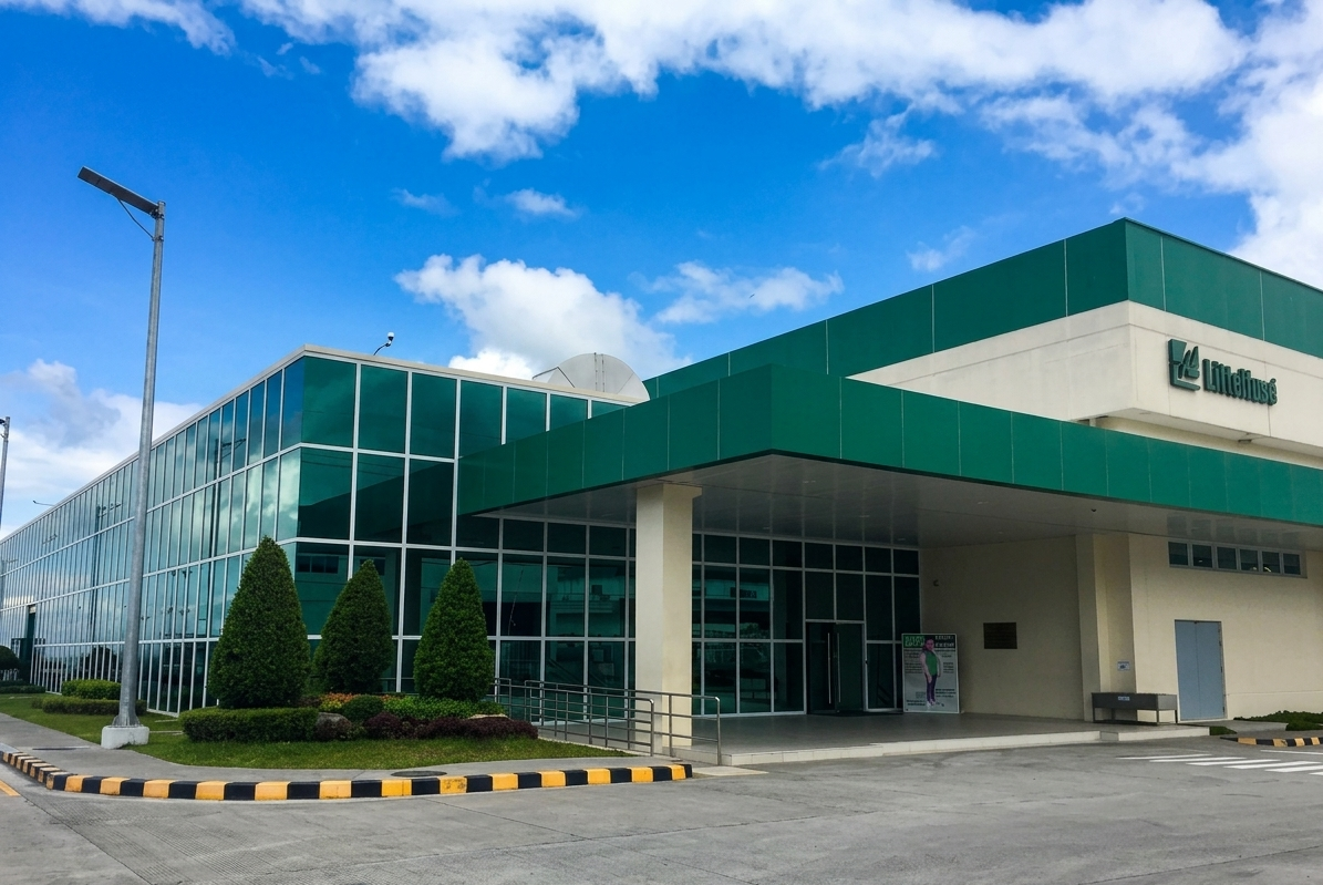 Exterior of a building with green and white facade, large glass windows, trees, and a parking lot against a partly cloudy sky.