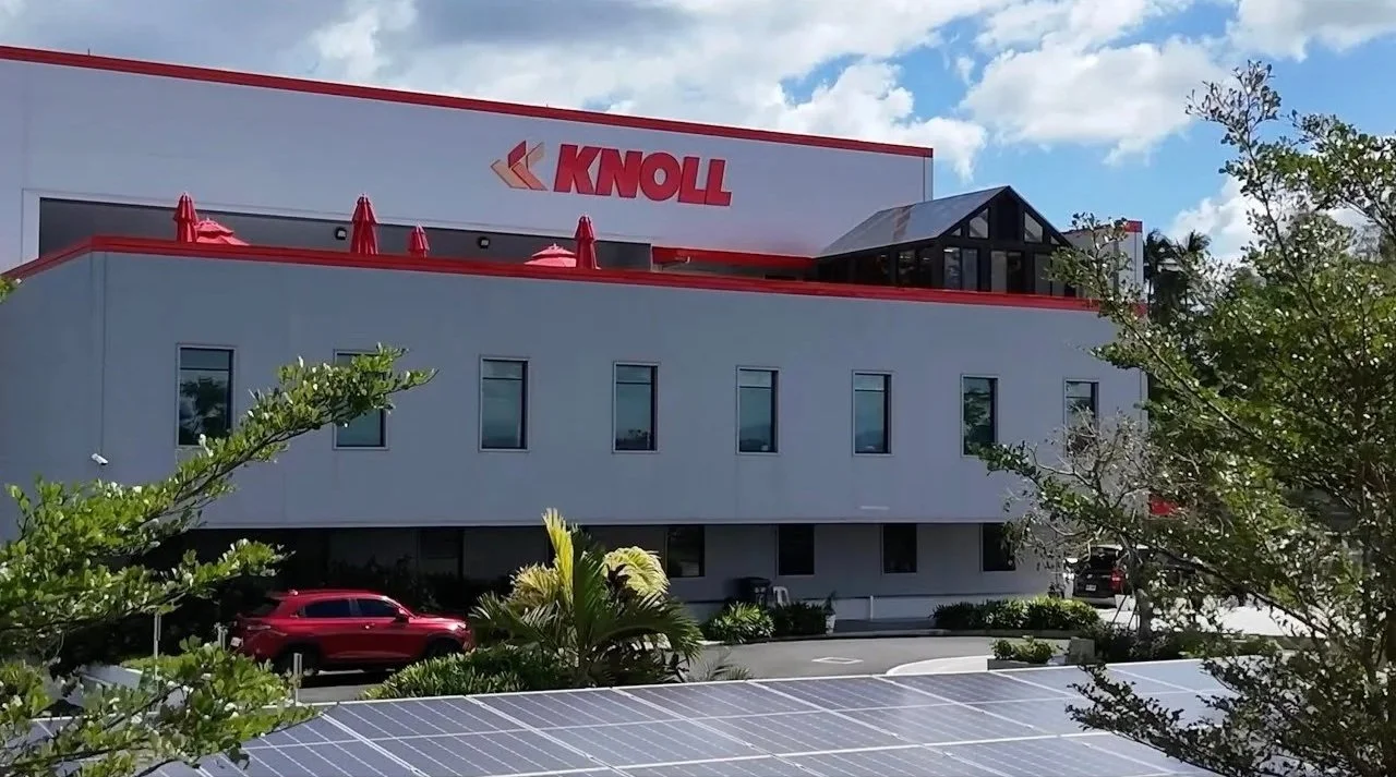 A commercial building with a large sign that says 'KNOLL' on the top. The building has a red and gray color scheme, with red umbrellas on a rooftop terrace, several windows, and cars parked in front. There are solar panels in the foreground, trees, and a partly cloudy sky.
