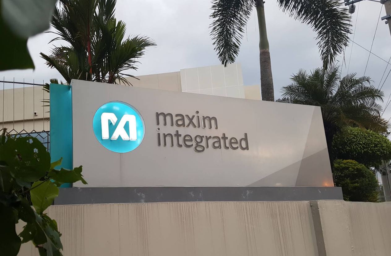 Sign for Maxim Integrated company with logo and company name against a backdrop of palm trees and cloudy sky.