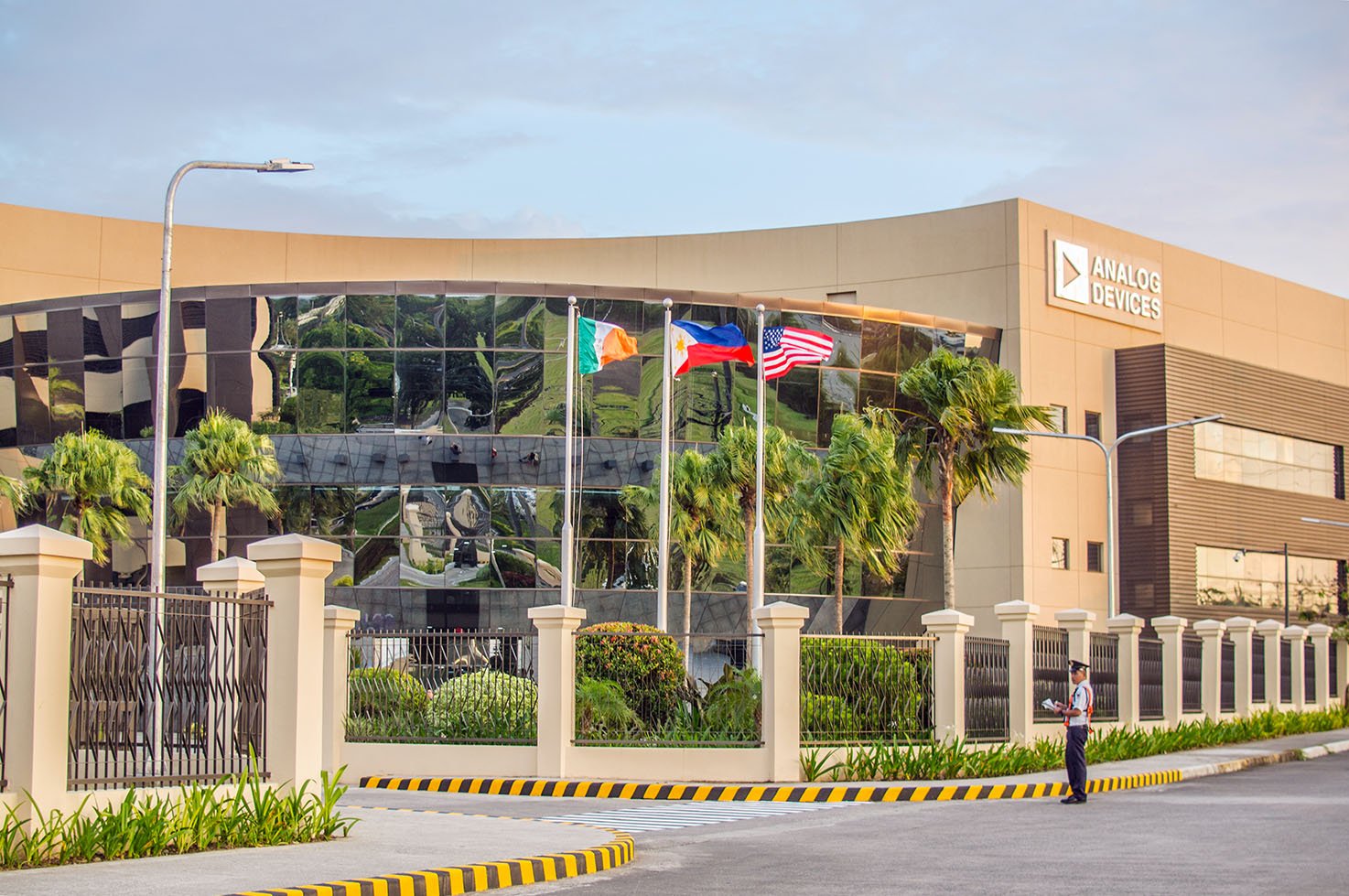 Exterior of the Analog Devices building with flags, trees, a security guard, and a fence.
