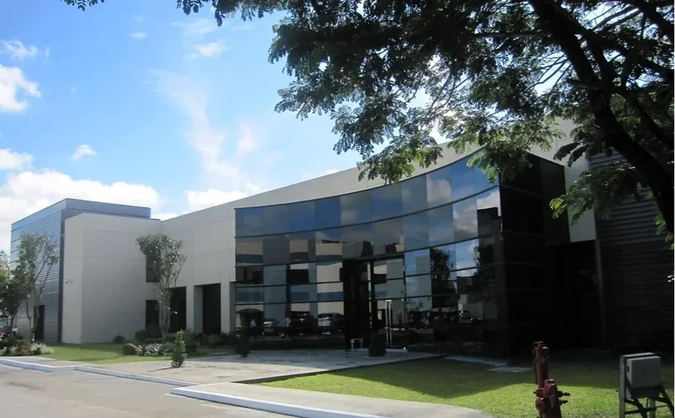 Modern commercial building with reflective glass facade, surrounded by trees and a parking lot under a partly cloudy sky.
