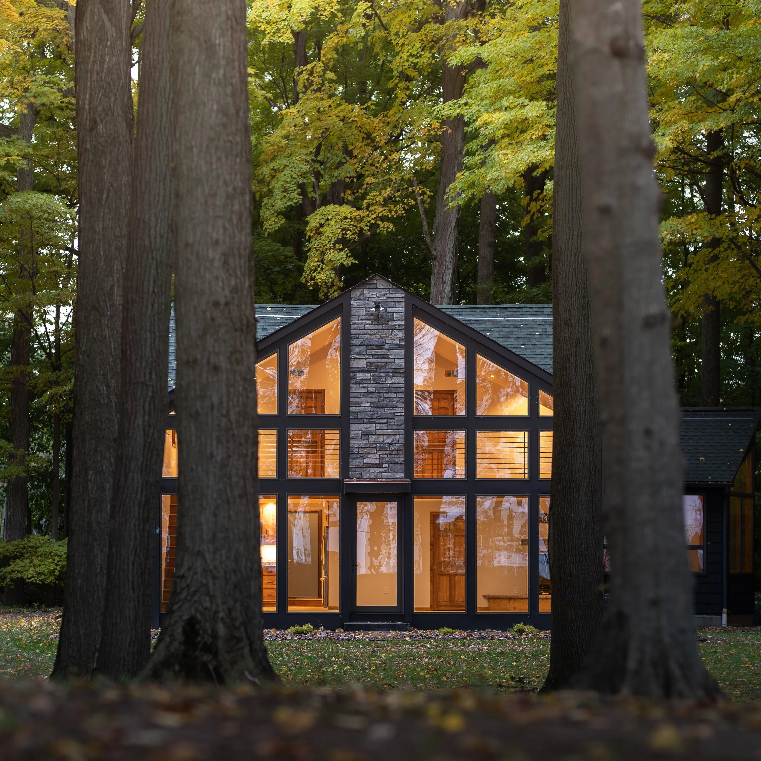 Modern house with large glass windows and stone accents, surrounded by tall trees in a wooded area, illuminated from inside during dusk or evening.