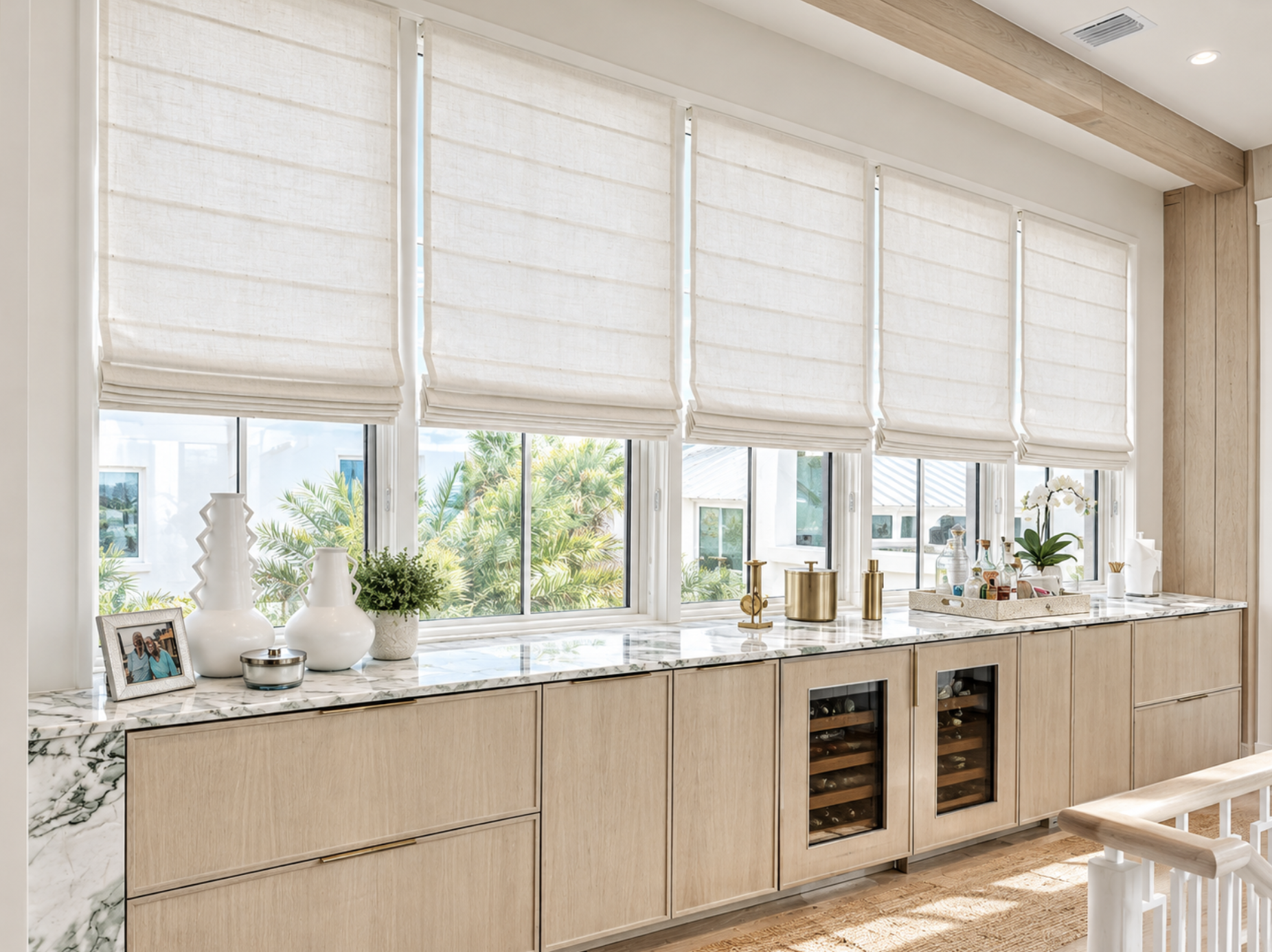 Sunlight coming through beige Roman shades covering a large window in a modern kitchen with light wood cabinetry and a marble countertop decorated with vases, photo frame, and small accessories.