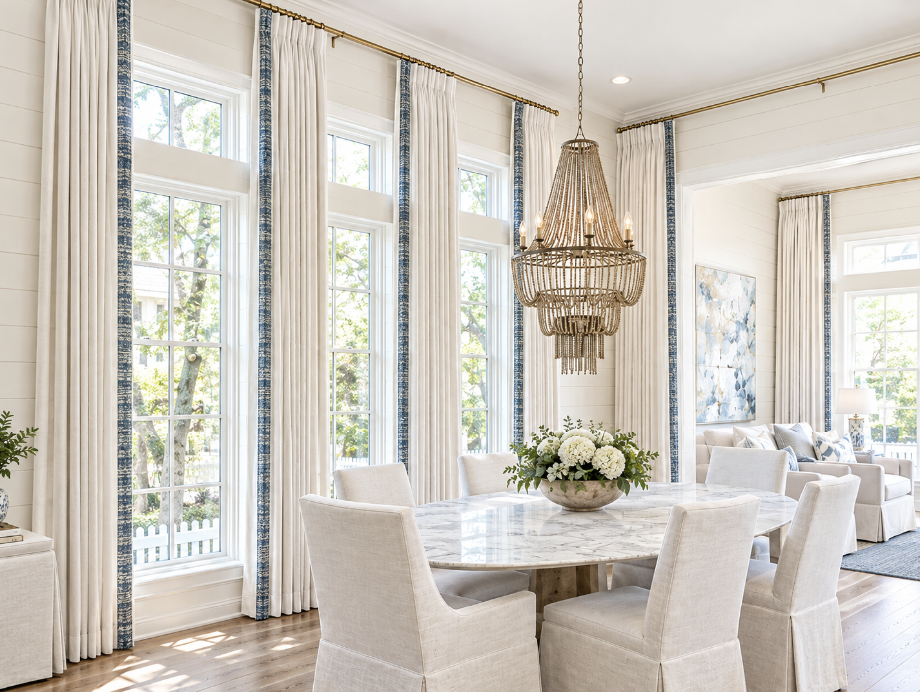 A bright, airy dining room with large windows, white curtains with blue accents, a chandelier, a marble dining table with cream chairs, and a floral centerpiece.