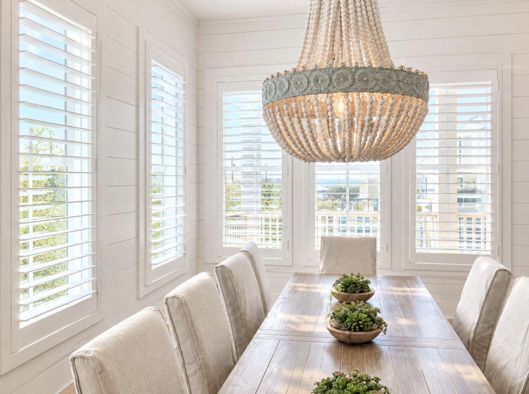 A dining room with large windows with white plantation shutters, beige upholstered chairs around a wooden table, a large beaded chandelier hanging from the ceiling, and potted succulents as table decor.