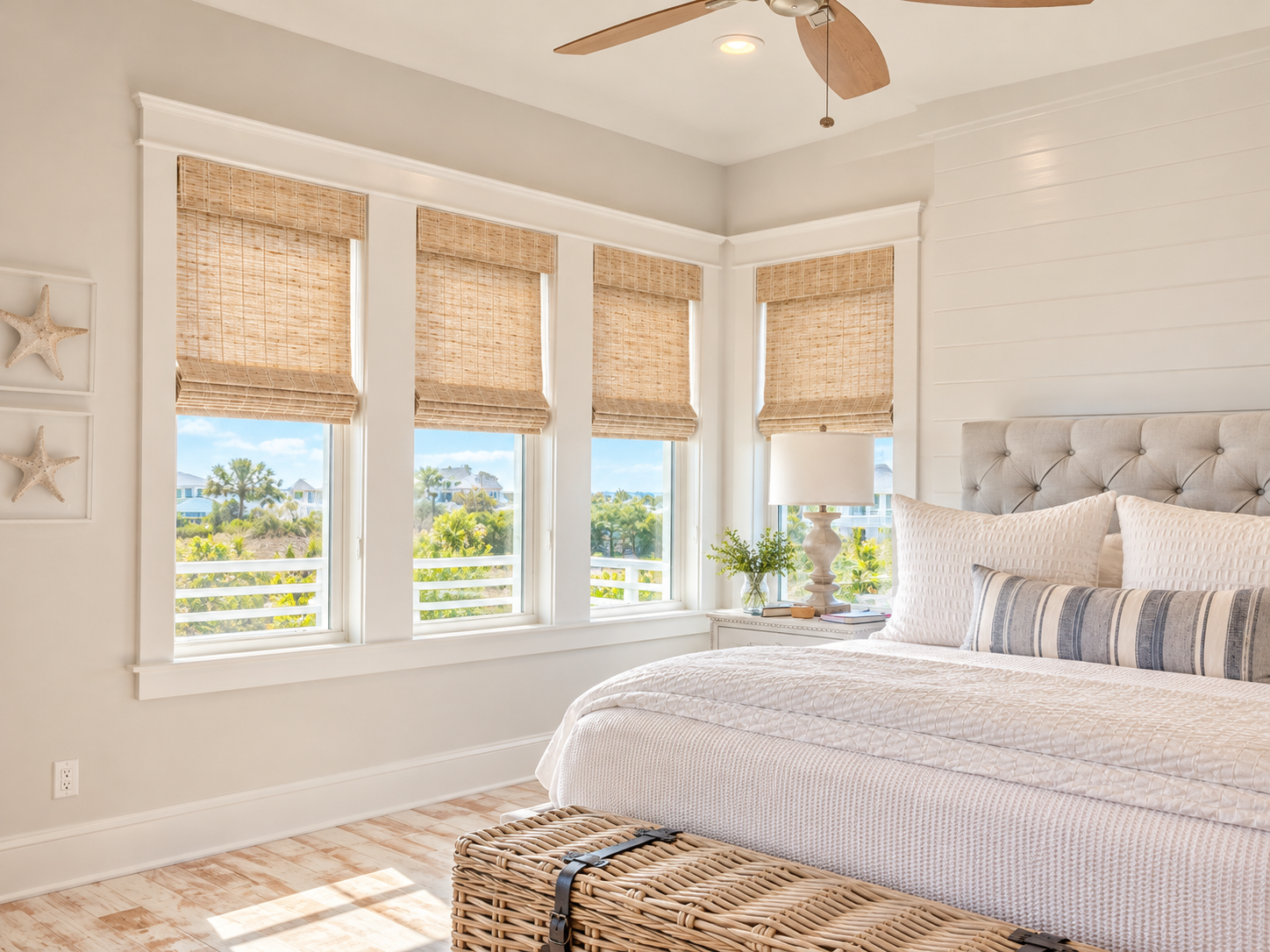 Bright bedroom with four large windows featuring woven blinds, white walls, a beige upholstered headboard, white bedding with multiple pillows, a white nightstand with a lamp and decorative items, and a woven rattan basket at the foot of the bed. Outside the windows, a view of trees and houses is visible.