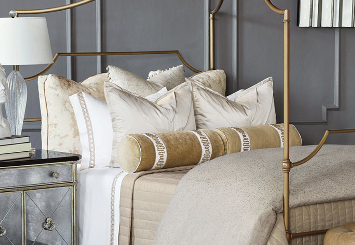A luxurious bed with gold-colored metal frame, decorated with cream and beige pillows and a beige bedspread, in a modern bedroom with gray paneled walls.