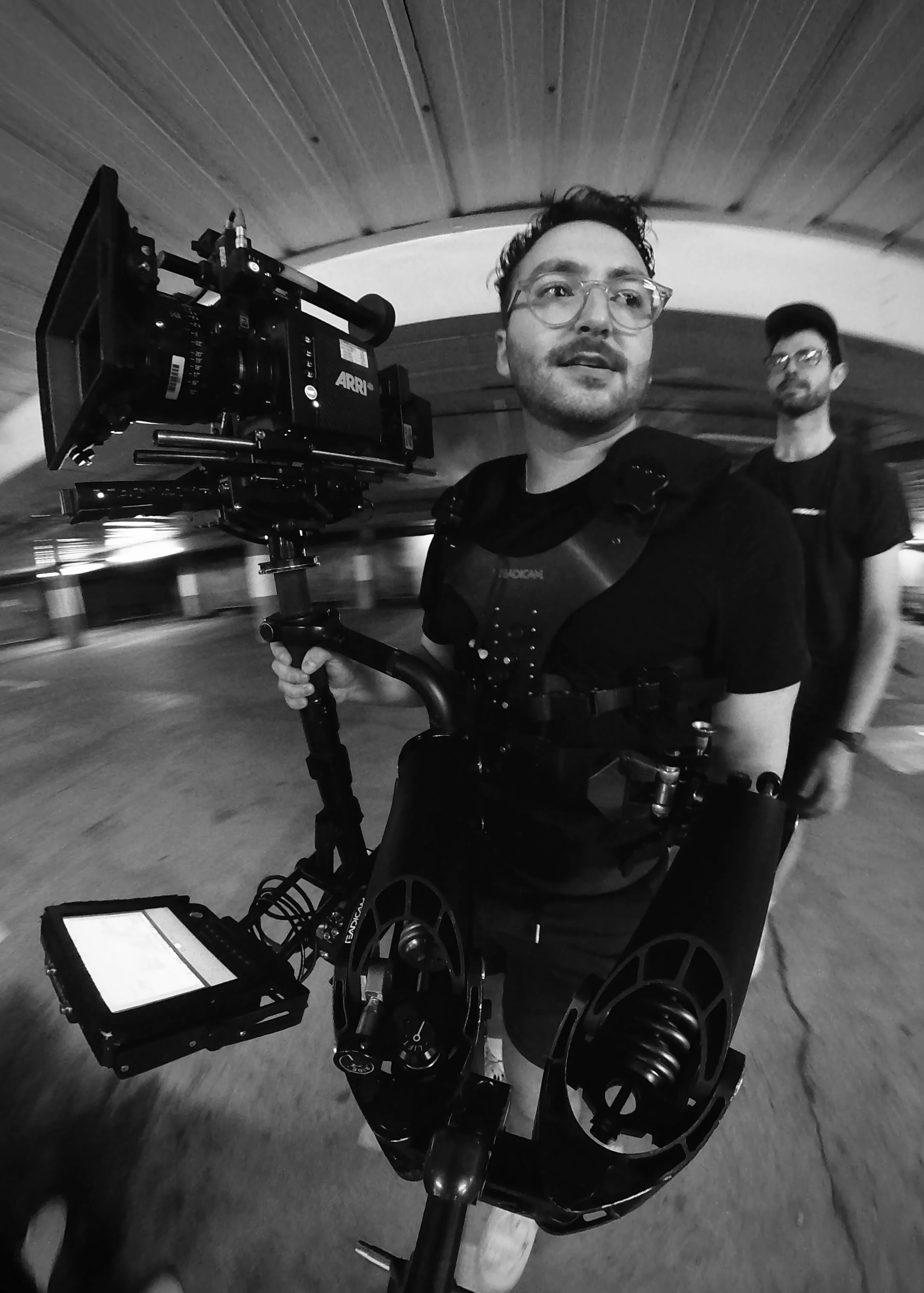 A person operating a professional camera mounted on a stabilised rig, with another person in the background, in a parking garage.