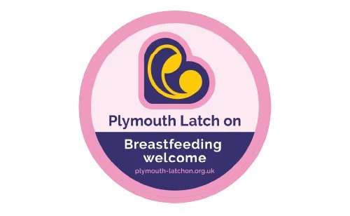 Pink and purple circular badge with text and a logo promoting breastfeeding support at Plymouth Latch on. The logo features a yellow moon and circle inside a dark purple heart outline. The text reads "Plymouth Latch on Breastfeeding welcome" and includes the website "plymouth-latchon.org.uk."
