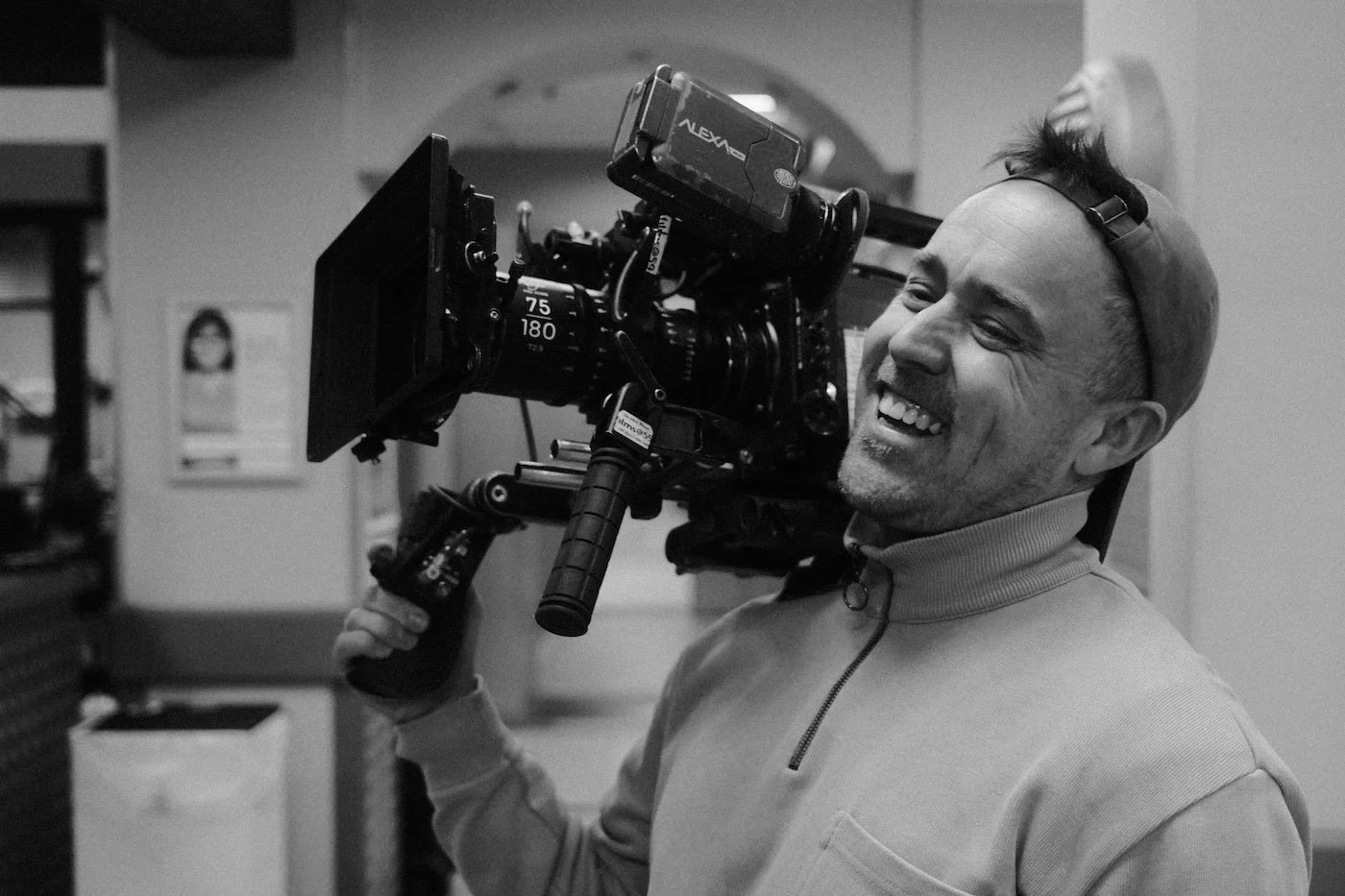 A man smiling and holding a professional camera rig on his shoulder, wearing a cap backwards and a zip-up jacket.