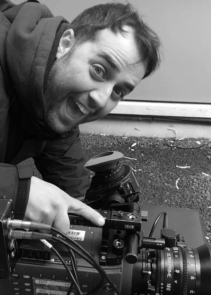 A man smiling excitedly while operating a professional video camera outdoors on the ground.
