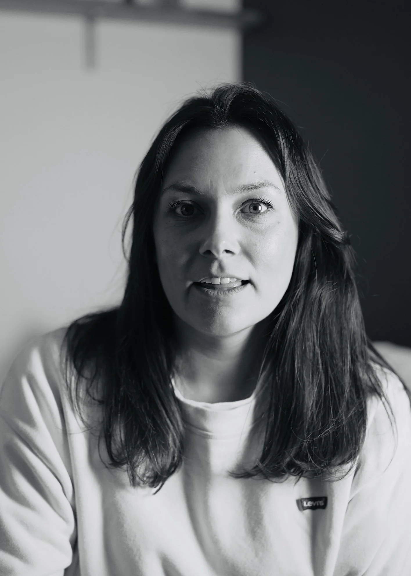 A black-and-white photo of a woman with medium-length dark hair, wearing a light-colored Levi's sweatshirt, looking directly at the camera with a neutral expression.