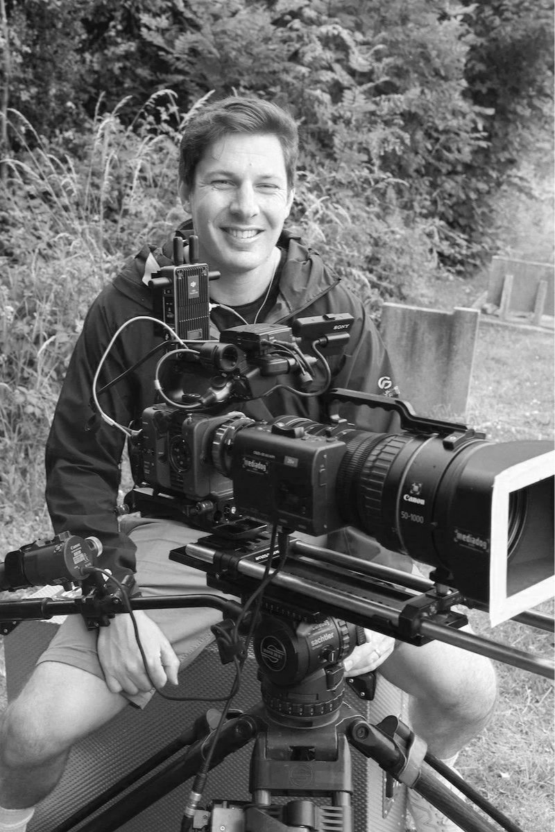 A man operating a professional video camera outdoors, with trees and foliage in the background, smiling at the camera.