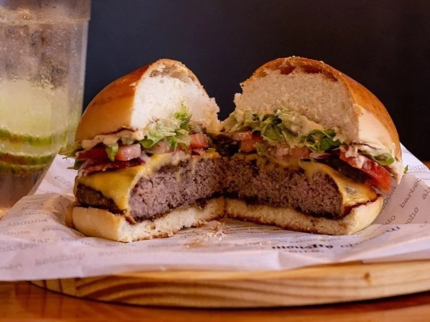 Half of a cheeseburger with lettuce, tomato, cheese, and beef patty, served on a bun with a side of a drink with lime slices.
