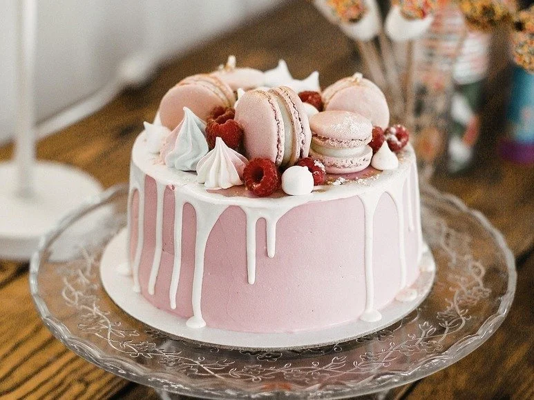A pink cake with white drip icing, decorated with pink macarons, raspberries, white meringue kisses, and small dollops of whipped cream, set on a glass cake stand.