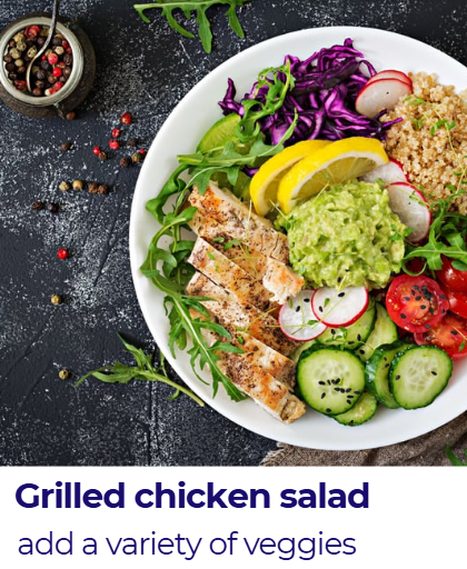 A white plate of grilled chicken salad with mixed greens, sliced cucumbers, radishes, cherry tomatoes, purple cabbage, lemon wedges, guacamole, and quinoa, garnished with herbs. A small bowl of mixed peppercorns is visible nearby.