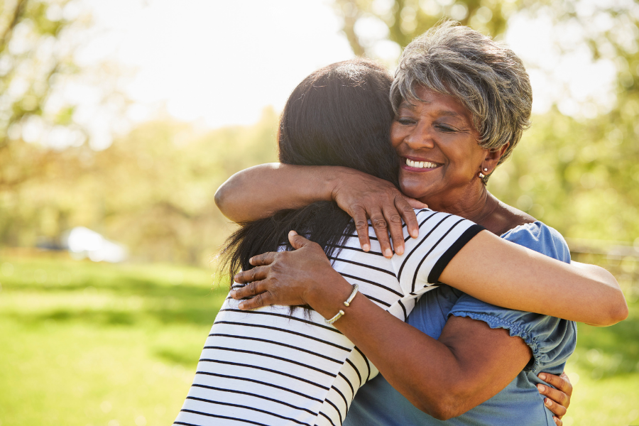 Two women hugging outdoors in a park on a sunny day, smiling warmly at each other.